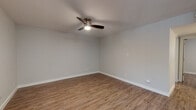 This single-floor unit features a clean, modern interior with consistent wood-look luxury vinyl plank flooring throughout, complemented by neutral gray walls and crisp white baseboards. The open layout is clearly visible in the floor plan view, revealing a spacious main living area that flows toward the kitchen and secondary rooms.

The kitchen is well-appointed with white shaker-style cabinetry, a stone-look countertop peninsula with an integrated sink, a decorative tile backsplash, and a full suite of stainless steel appliances including a gas range, microwave, and refrigerator. Recessed lighting illuminates the workspace effectively.

The bedrooms are generously sized, each featuring ceiling fans with integrated lighting and neutral painted walls that lend a bright, airy feel. One bedroom benefits from a window with vertical blinds, allowing natural light to filter in. A panel-style entry door with a brushed nickel lockset is visible from the main living area.

The bathroom showcases large-format marble-look wall tile surrounding the tub and shower combination, complete with a built-in niche, brushed nickel fixtures, and a grab bar. Wood-look flooring continues into the bathroom, maintaining design cohesion throughout the unit.