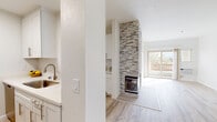 This thoughtfully updated single-floor residence showcases a clean, modern aesthetic throughout, highlighted by light-toned hardwood-style flooring that flows seamlessly from room to room. The open living area serves as the heart of the space, anchored by a striking floor-to-ceiling stacked stone fireplace with a sleek glass front — a standout architectural feature that adds warmth and visual drama. A ceiling fan overhead and a sliding glass door opening to a private balcony with outdoor views enhance the room's airy, comfortable feel. The kitchen is well-appointed with white shaker cabinetry, quartz countertops, a white subway tile backsplash, and a full suite of stainless steel appliances including a refrigerator, electric range, and over-the-range microwave. A raised breakfast bar connects the kitchen to the living area, promoting an open and social layout. The bedroom offers a tranquil retreat with a large window framing tree-lined outdoor views, a baseboard heater, and a mirrored sliding closet. The bathroom is clean and functional, featuring a combined tub and shower, white vanity with brushed nickel hardware, and matching light flooring. Throughout the space, bright white walls and abundant natural light contribute to a fresh, inviting atmosphere.