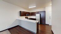 Located at 1151 32nd Ave N in Fargo, North Dakota, this single-floor unit offers a clean, functional layout designed for comfortable everyday living. The kitchen serves as a central highlight, featuring rich dark wood cabinetry, white countertops, stainless steel appliances including a refrigerator, electric range, microwave, and dishwasher, all complemented by durable hardwood-style flooring. A peninsula counter extends into the adjacent living area, creating an open, connected feel between cooking and living spaces. The living area is carpeted and receives natural light through a large window, contributing to a bright and welcoming atmosphere. Multiple bedrooms are found throughout the unit, each finished with plush carpet, neutral walls, and windows fitted with white blinds that allow for natural light while maintaining privacy. The bathroom is thoughtfully designed, offering a spacious layout with a dedicated vanity area featuring dark wood cabinetry, a wide mirror, and a tall storage tower. A separate water closet houses the toilet, adding a functional separation of space. Throughout the unit, dark baseboards provide a clean contrast to the light-toned walls, lending a cohesive and modern aesthetic to the overall interior design.