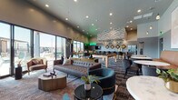 This contemporary community clubroom is thoughtfully designed to serve as a dynamic social hub, blending entertainment, relaxation, and modern aesthetics in a single open-plan space. The layout is anchored by a generous lounge area furnished with tufted leather sofas, velvet accent chairs, and layered patterned carpeting that adds depth and texture throughout. A dramatic floor-to-ceiling textured stone fireplace anchors one wall, paired with a wall-mounted television and curated sculptural décor, creating an inviting focal point for casual gatherings. Adjacent to the lounge, a dedicated game zone features a full-size billiards table illuminated by pendant lighting, with a built-in banquette seating area accented by a glowing neon sign, adding a playful, energetic vibe. The wet bar area stands out with its striking geometric 3D ceiling treatment, pendant globe lights, tufted bar stools, and a full-size refrigerator, making it ideal for hosting. Floor-to-ceiling glass walls and sliding doors flood the space with natural light and provide direct sightlines to an outdoor terrace area. High-top bistro tables positioned near the windows offer additional casual seating with exterior views. Neon art installations and bold wall décor lend a modern, vibrant character to the entire space.