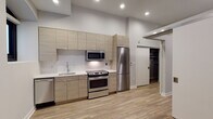 This single-floor space showcases a clean, contemporary design with consistent wood-look flooring and neutral tones carried throughout. The kitchen serves as a standout feature, equipped with a full suite of stainless steel appliances including a refrigerator, electric range, built-in microwave, and dishwasher. Light wood flat-panel cabinetry extends to the ceiling, maximizing storage, while white subway tile backsplash and light quartz countertops contribute to the bright, modern aesthetic. An arched window in the kitchen provides natural light and an architectural detail that adds character to the space. A hallway connects the main living areas to the additional rooms, lined with recessed lighting and featuring a walk-in closet with wire shelving for organized storage. A bedroom with a sliding barn-style door offers a minimalist layout with angled ceiling detail, adding subtle architectural interest. The bathroom is finished with a frameless glass-enclosed shower, wood-look tile flooring, and a floating vanity with a clean white countertop, maintaining the cohesive modern aesthetic found throughout the unit. Recessed lighting is used consistently throughout, enhancing the bright, polished ambiance of the entire space.