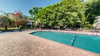 Located at 800 State St in West Columbia, South Carolina, this outdoor pool area offers a well-appointed communal retreat designed for relaxation and recreation. The centerpiece is a generously sized swimming pool with clear turquoise water, accessible via stainless steel entry rails and marked with clearly visible depth indicators. The pool is surrounded by an expansive brick paver deck that provides ample space for lounging and socializing.

Scattered throughout the deck are multiple lounge chairs and seating arrangements, including umbrella-shaded tables that offer relief from the South Carolina sun. The entire pool area is enclosed by a wrought iron security fence, ensuring a safe and private environment. A low-profile brick building serves as the pool house, providing additional facilities for guests.

The surrounding landscape is a defining feature of this space, with towering mature hardwood trees and lush Southern vegetation — including striking palm trees and flowering crepe myrtles — creating a verdant, canopy-like backdrop that lends the area a shaded, park-like atmosphere. The combination of classic brick architecture, manicured greenery, and thoughtfully arranged outdoor furnishings makes this pool area a comfortable and visually appealing outdoor amenity.