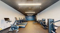 Located at 1155 S Rangeline Rd in Carmel, Indiana, this fitness facility offers a single-floor exercise room designed to support a full range of workout routines. The space features a sleek, modern aesthetic defined by dark drop-tile ceilings with recessed lighting and bright linear LED fixtures that provide strong, even illumination throughout. The walls are finished in clean white, accented by bold teal-blue panels that add a contemporary visual character to the interior.

The exercise room is generously proportioned, providing ample space for both cardio and strength training areas. Cardio equipment includes Matrix treadmills, stationary bikes, and ellipticals, all arranged in an organized row along one side. The strength training section features a multi-function cable machine, a power rack with free weights, dumbbell racks with benches, and additional resistance machines from brands including Hoist and Precor. Durable rubberized flooring covers the workout area, offering safety and stability underfoot.

Natural light filters into the space through large glass double-entry doors facing the street, offering views of the surrounding outdoor environment. The layout flows logically from the entry point through the cardio zone and into the strength area, making navigation intuitive. Overall, the facility presents a professional and functional environment well-suited for comprehensive fitness training.
