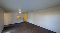 This single-floor residential unit offers a straightforward and functional layout designed for comfortable everyday living. The main living area is generously sized, featuring dark wall-to-wall carpeting that anchors the space and provides a warm underfoot feel. A textured popcorn ceiling spans the room overhead, fitted with a ceiling fan that promotes air circulation throughout. The walls are painted in a clean, neutral white, creating a bright and open atmosphere that is easy to personalize.

A distinctive pass-through window and counter connects the living area to the kitchen, which is accented with warm yellow walls and overhead cabinetry. This feature enhances the sense of openness between the two spaces while maintaining a clear functional separation. The kitchen is visible through both the pass-through and a connecting doorway, offering convenient access from the main living area.

A sliding mirrored closet is visible near the entry, adding both practical storage and a reflective element that helps to visually expand the space. A hallway leads further into the unit, suggesting additional rooms beyond the main living zone. Natural light filters in through at least one window, casting a soft glow across the carpeted floor. Overall, the unit presents a clean, move-in ready canvas with a cohesive, understated aesthetic throughout.