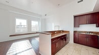 Located in the heart of downtown Los Angeles, this one-bedroom, one-bathroom unit at 215 W 5th Street offers a well-appointed layout across a single floor. Rich, warm-toned hardwood flooring runs throughout the living and bedroom areas, complemented by crisp white walls that amplify the natural light pouring in through multiple windows.

The open-concept living area flows seamlessly into the kitchen, which features deep mahogany cabinetry, granite countertops, stainless steel appliances including a refrigerator and microwave, a built-in dishwasher, and a peninsula that doubles as a breakfast bar. The kitchen transitions to light-toned tile flooring, clearly delineating the space.

The living room is generously proportioned with soaring ceilings and a built-in window bench framing dual windows that offer views of the surrounding downtown Los Angeles skyline, flooding the room with natural light throughout the day.

The bedroom is a comfortable, well-sized retreat with the same warm hardwood floors and ample wall space. The bathroom is finished with large-format cream marble tile, a soaking tub with a glass surround, a mahogany vanity with granite countertop, and elegant chrome fixtures. A hallway connects the rooms, providing a practical and efficient flow throughout the unit.