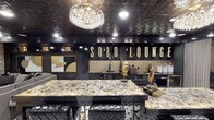 SoBo Lounge is a meticulously designed single-floor entertainment venue that exudes luxury through a bold black-and-gold design language carried consistently throughout the space. The centerpiece of the lounge is a striking bar area anchored by a dramatically veined marble-top island with tufted black leather bar seating. Behind the bar, illuminated marquee-style lettering spells out "SoBo Lounge" against a gold textured backsplash, while dark cabinetry and a full draft beer setup underscore its functionality. A grand crystal chandelier overhead adds glamour, complemented by an ornate embossed tin ceiling finished in high-gloss black that enhances the dramatic ambiance throughout the main area. The flooring features classic black-and-white hexagonal tile, adding a timeless yet modern foundation to the space. Adjacent to the bar, a relaxed lounge area offers plush grey sectional seating, a mirrored gold-framed coffee table, wall-mounted flat-screen TV, and curated gold-and-black artwork. Textured stone accent walls lend warmth and dimension to this retreat. A cozy built-in banquette nook with cushioned seating and decorative pillows provides an intimate gathering spot. A separate room features matching stone walls, elegant fringe wall sconces, gold-toned accent furnishings, and thoughtfully placed artwork, making it ideal as a private lounge or VIP area.
