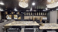 SoBo Lounge is a meticulously designed single-floor entertainment venue that exudes luxury through a bold black-and-gold design language carried consistently throughout the space. The centerpiece of the lounge is a striking bar area anchored by a dramatically veined marble-top island with tufted black leather bar seating. Behind the bar, illuminated marquee-style lettering spells out "SoBo Lounge" against a gold textured backsplash, while dark cabinetry and a full draft beer setup underscore its functionality. A grand crystal chandelier overhead adds glamour, complemented by an ornate embossed tin ceiling finished in high-gloss black that enhances the dramatic ambiance throughout the main area. The flooring features classic black-and-white hexagonal tile, adding a timeless yet modern foundation to the space. Adjacent to the bar, a relaxed lounge area offers plush grey sectional seating, a mirrored gold-framed coffee table, wall-mounted flat-screen TV, and curated gold-and-black artwork. Textured stone accent walls lend warmth and dimension to this retreat. A cozy built-in banquette nook with cushioned seating and decorative pillows provides an intimate gathering spot. A separate room features matching stone walls, elegant fringe wall sconces, gold-toned accent furnishings, and thoughtfully placed artwork, making it ideal as a private lounge or VIP area.