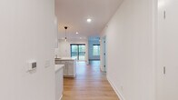 This single-floor space showcases a clean, modern design highlighted by light-toned hardwood flooring, crisp white walls, and recessed lighting throughout. The open-concept layout seamlessly connects the kitchen and living area, creating a cohesive and functional flow.

The kitchen is thoughtfully designed with white shaker-style cabinetry extending to the ceiling, offering ample storage. Bright white quartz countertops run along the perimeter and wrap around a peninsula with an integrated sink, providing generous workspace and casual seating potential. Stainless steel appliances — including a range with electric cooktop, over-the-range microwave, refrigerator, and dishwasher — complement the contemporary aesthetic. Pendant lighting with Edison-style bulbs adds a warm, decorative touch above the peninsula.

The living area is spacious and filled with natural light, courtesy of large black-framed windows and a sliding glass door that opens to a private balcony overlooking lush, tree-lined surroundings. Recessed ceiling lights and a ceiling fan ensure a comfortable ambiance year-round.

The bathroom features a walk-in shower with sliding glass doors, a white vanity with an integrated sink, and light wood-look flooring, maintaining the space's consistent, polished aesthetic throughout.