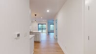 This single-floor space showcases a clean, modern design highlighted by light-toned hardwood flooring, crisp white walls, and recessed lighting throughout. The open-concept layout seamlessly connects the kitchen and living area, creating a cohesive and functional flow.

The kitchen is thoughtfully designed with white shaker-style cabinetry extending to the ceiling, offering ample storage. Bright white quartz countertops run along the perimeter and wrap around a peninsula with an integrated sink, providing generous workspace and casual seating potential. Stainless steel appliances — including a range with electric cooktop, over-the-range microwave, refrigerator, and dishwasher — complement the contemporary aesthetic. Pendant lighting with Edison-style bulbs adds a warm, decorative touch above the peninsula.

The living area is spacious and filled with natural light, courtesy of large black-framed windows and a sliding glass door that opens to a private balcony overlooking lush, tree-lined surroundings. Recessed ceiling lights and a ceiling fan ensure a comfortable ambiance year-round.

The bathroom features a walk-in shower with sliding glass doors, a white vanity with an integrated sink, and light wood-look flooring, maintaining the space's consistent, polished aesthetic throughout.