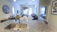This single-floor residence offers a seamlessly connected open-concept layout that integrates the kitchen, living area, and bedroom into one cohesive living environment. The kitchen serves as the focal point of daily life, showcasing polished granite countertops and a sleek stainless steel sink with a high-arc pull-down faucet — a combination that delivers both aesthetic appeal and everyday functionality.

The kitchen flows effortlessly into the living area, which is generously sized and filled with natural light streaming through large windows dressed with elegant draping curtains. A ceiling fan overhead ensures comfortable airflow throughout the space. The living area is well-suited for both relaxation and entertaining, with ample room for a full sectional sofa, layered area rugs, and accent lighting provided by a striking arc floor lamp with a dome shade.

A peek into the bedroom reveals a bright, airy retreat with windows that invite natural light, creating a calm and restful atmosphere. Throughout the home, the light-toned walls and cohesive design choices lend the space a clean, contemporary feel. Overall, this residence balances modern finishes with a warm, livable ambiance that makes it both functional and inviting.