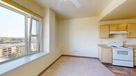 This single-floor unit offers a well-organized layout comprising a living room, kitchen, bathroom, and closet. The living room is finished with plush neutral carpet, warm cream-toned walls, and a ceiling fan overhead, creating a comfortable and airy atmosphere. A large window floods the space with natural light and frames elevated views of the surrounding cityscape and rooftops, adding a notable sense of openness to the compact footprint.

The kitchen transitions seamlessly from the living area, distinguished by wood-look plank flooring that contrasts neatly with the carpeted living space. Light maple cabinetry lines the walls, providing ample storage above and below the countertops. The kitchen is equipped with a white electric range, a dishwasher, a refrigerator, and a single-basin sink — offering a fully functional cooking environment. Under-cabinet range hood ventilation and a clean countertop workspace complete the practical setup.

The closet features a sliding door entry with an interior hanging rod, offering dedicated storage within the unit. Throughout the space, consistent warm neutral wall tones, wood-trimmed baseboards, and ceiling fans contribute to a cohesive and functional interior aesthetic suited for efficient everyday living.