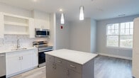 This single-floor unit in East Providence, Rhode Island, presents a thoughtfully designed interior with a contemporary aesthetic throughout. The open-concept kitchen serves as the centerpiece of the living space, outfitted with white upper cabinetry, a mosaic tile backsplash, and a full suite of stainless steel appliances including a range, over-the-range microwave, refrigerator, and dishwasher. A two-toned center island with a crisp white countertop and contrasting blue-gray base provides additional workspace and storage, illuminated by sleek pendant lighting overhead. Light wood-tone luxury vinyl plank flooring extends seamlessly across the entire unit, enhancing the sense of continuity and openness. A stacked washer and dryer unit is conveniently integrated into the kitchen area, adding practical in-unit laundry. The bedroom offers a calm, neutral palette with soft gray walls, natural light from a double-hung window with an outdoor view, and a generously sized closet with built-in shelving. The bathroom features wood-look tile flooring, a white vanity with a clean countertop, and a full tub and shower combination. Recessed and pendant lighting throughout maintains a bright, welcoming ambiance across the unit.