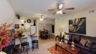 This single-story residence at 1800 Links Blvd in Tuscaloosa, Alabama, offers an open-concept floor plan that seamlessly connects the main living areas. The living room is a comfortable and well-appointed space, featuring carpet flooring, a ceiling fan with light fixture, and room for substantial furniture arrangements. A bold wall art piece and warm lamp lighting contribute to the welcoming ambiance. Adjacent to the living area, the dining space accommodates a full dining table and chairs, and shares the same open layout, making it ideal for entertaining. Ceiling fans are present in both the living and dining areas, promoting airflow throughout. The kitchen is accessible via a breakfast bar with bar stool seating, providing a casual dining option. White cabinetry and white appliances give the kitchen a clean, classic look, while a sink with a window above it adds functional convenience. The transition between the kitchen's tile-style flooring and the carpeted living area clearly defines each zone within the open layout. Overall, the space presents a cohesive, functional design with a warm and livable atmosphere.