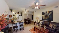 This single-story residence at 1800 Links Blvd in Tuscaloosa, Alabama, offers an open-concept floor plan that seamlessly connects the main living areas. The living room is a comfortable and well-appointed space, featuring carpet flooring, a ceiling fan with light fixture, and room for substantial furniture arrangements. A bold wall art piece and warm lamp lighting contribute to the welcoming ambiance. Adjacent to the living area, the dining space accommodates a full dining table and chairs, and shares the same open layout, making it ideal for entertaining. Ceiling fans are present in both the living and dining areas, promoting airflow throughout. The kitchen is accessible via a breakfast bar with bar stool seating, providing a casual dining option. White cabinetry and white appliances give the kitchen a clean, classic look, while a sink with a window above it adds functional convenience. The transition between the kitchen's tile-style flooring and the carpeted living area clearly defines each zone within the open layout. Overall, the space presents a cohesive, functional design with a warm and livable atmosphere.