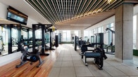 Located in Portland's vibrant lower Burnside neighborhood, this expansive fitness center is a well-appointed facility designed to support a wide range of workout routines. The space is defined by an architectural slatted wood ceiling with integrated linear lighting that lends a refined, contemporary aesthetic throughout.

Large floor-to-ceiling windows line the perimeter, flooding the facility with natural light and offering impressive panoramic views of the Portland skyline and surrounding cityscape — a standout feature that sets the tone for the entire space.

The fitness floor is divided into distinct functional zones. A strength training area features a comprehensive selection of resistance machines, free weights, adjustable benches, and a full dumbbell rack, complemented by wall-to-wall mirrors and wall-mounted TVs. A dedicated functional training zone offers TRX suspension systems, a heavy punching bag, and air bikes on warm hardwood flooring. The cardio section is equipped with treadmills, Matrix stair climbers, rowing machines, ellipticals, and connected stationary cycles. A separate studio area with light hardwood floors, exposed concrete columns, and ample open space is outfitted with medicine balls, aerobic step platforms, and resistance accessories — ideal for group or solo training sessions. Rubber flooring throughout the main workout areas ensures both safety and durability.