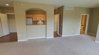 This single-floor residence offers a straightforward and functional layout designed to maximize usability. The living area is finished with wall-to-wall carpet in a neutral tone, providing a comfortable and cohesive feel. Walls are painted in a warm beige, creating a consistent palette throughout the visible spaces.

A defining architectural feature is the arched pass-through opening that connects the living area to the kitchen, adding a classic design element while maintaining an open sightline. The kitchen is equipped with light wood cabinetry, a white electric range, an over-the-range microwave, and a refrigerator. The kitchen counter extends slightly to form a bar-style ledge, offering casual dining or prep space.

Ceiling-mounted HVAC vents are present in both the living and kitchen areas, ensuring climate control throughout. A hallway visible beyond the living space transitions to hardwood or laminate flooring, leading toward additional rooms. The overall ambiance is clean, simple, and move-in ready, suited for practical everyday living.