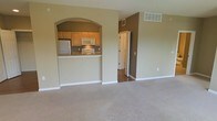 This single-floor residence offers a straightforward and functional layout designed to maximize usability. The living area is finished with wall-to-wall carpet in a neutral tone, providing a comfortable and cohesive feel. Walls are painted in a warm beige, creating a consistent palette throughout the visible spaces.

A defining architectural feature is the arched pass-through opening that connects the living area to the kitchen, adding a classic design element while maintaining an open sightline. The kitchen is equipped with light wood cabinetry, a white electric range, an over-the-range microwave, and a refrigerator. The kitchen counter extends slightly to form a bar-style ledge, offering casual dining or prep space.

Ceiling-mounted HVAC vents are present in both the living and kitchen areas, ensuring climate control throughout. A hallway visible beyond the living space transitions to hardwood or laminate flooring, leading toward additional rooms. The overall ambiance is clean, simple, and move-in ready, suited for practical everyday living.