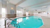 Located at 1275-1285 Richmond Rd in Ottawa, Ontario, this indoor aquatic facility offers a generously sized, single-floor space designed for both recreational swimming and lap training. The centerpiece is a large rectangular lap pool with clearly marked lanes, featuring stainless steel entry rails and underwater lighting that gives the water a vibrant turquoise appearance. A separate, rounded hot tub area is positioned at one end of the facility, providing a dedicated space for relaxation and hydrotherapy.

The architecture is characterized by high, vaulted ceilings with exposed beams and a combination of fluorescent strip lighting and wall-mounted sconces, ensuring the space is well-lit throughout. Large windows along one wall allow natural light to filter in and offer outdoor views, enhancing the open, airy ambiance of the room. The walls are finished with a neutral tile wainscoting, complemented by white paneling above, creating a clean and functional aesthetic.

The poolside deck is finished in small-format mosaic tiles, providing slip-resistant surfaces around the pools. Stepped seating areas with aggregate finishes offer spaces for observers or resting swimmers. Safety equipment, including life rings and posted pool rules in both English and French, is prominently displayed throughout. A separate entry and change room area is accessible via steps near the main entrance of the facility.