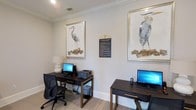 Located at 28013 Hwy 181 in Daphne, Alabama, this business center offers a focused and professional environment designed to meet productivity needs. The single-floor space is thoughtfully appointed with multiple dedicated computer workstations, each equipped with desktop computers and a shared printer, providing everything needed for work or administrative tasks. The room is finished with neutral, light-toned walls and crown molding, lending a clean and polished aesthetic throughout. Tasteful coastal-inspired artwork featuring elegant heron illustrations in gilded frames adorns the walls, adding a sophisticated decorative touch that complements the surrounding Gulf Coast region. Soft, recessed lighting and a table lamp contribute to a well-lit and comfortable atmosphere conducive to focused work. The light wood-toned flooring and dark wood desks create a balanced contrast that feels both modern and inviting. Ergonomic seating ensures comfort during extended use. Prominently displayed business center rules provide clear guidelines for users. Overall, this space strikes a balance between functional utility and refined design, making it a welcoming and efficient resource for those seeking a professional workspace.
