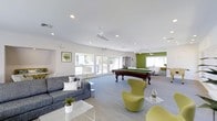 This single-floor community space is thoughtfully designed to encourage social interaction and leisure. The open-plan layout is filled with natural light streaming through large windows and glass doors that line the perimeter, creating an airy and welcoming atmosphere throughout.

The main lounge area features a contemporary gray sofa accented with decorative throw pillows, paired with distinctive egg-style chairs in a bold chartreuse yellow that add a playful, modern flair. A sleek glass coffee table anchors the seating arrangement, complemented by small potted plants that bring a touch of greenery indoors.

A full-size pool table serves as a central recreational focal point, positioned on plush carpeting that defines the activity zone. Ceiling fans are installed throughout, ensuring comfortable airflow across the entire space.

An accent wall painted in a vibrant olive green adds depth and visual interest to the far end of the room, where additional seating and open views through the windows create a secondary lounge area. The clean white walls and recessed lighting throughout enhance the bright, contemporary aesthetic.

Overall, the space strikes a balance between comfort and functionality, offering an inviting environment suited for both casual relaxation and group entertainment.