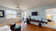 Located at 3201 Emerald Pointe Dr in Hollywood, Florida, this single-floor residence offers a well-organized layout with a cohesive, relaxed aesthetic throughout. Soft blue-gray walls and warm wood-style flooring run continuously through the main living areas, creating a calm, inviting atmosphere.

The living room is generously proportioned, featuring a ceiling fan, a textured ceiling, and multiple windows dressed with vertical blinds that allow ample natural light to filter in. A built-in wall-mounted media shelf provides a distinctive architectural detail, offering display space beneath a wall-mounted television. The room comfortably accommodates both a seating arrangement and a separate workspace or sitting area.

The kitchen is designed with an open peninsula that connects it to the living space, offering light wood cabinetry, white appliances, and a pendant light overhead. A dedicated laundry area with stacked washer and dryer units is conveniently located just off the kitchen.

The bedroom is a calm retreat, featuring the same soft wall tones, a textured ceiling, and vertical blinds covering a sliding door that provides access to an outdoor area. The bathroom is efficiently laid out with a single vanity, white cabinetry, a large mirror, and a shower-tub combination.