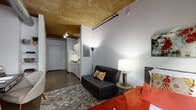 This single-floor studio showcases an industrial-loft aesthetic defined by exposed wood-plank ceilings with visible beams and track lighting overhead, paired with polished concrete floors throughout. The open layout connects the living and sleeping areas seamlessly, creating an efficient and cohesive living environment.

The sleeping area is anchored by a large, dark-framed window that draws in natural light while offering views of the surrounding urban exterior. A built-in wardrobe system with frosted glass sliding doors provides ample storage without interrupting the clean lines of the space. Exposed ductwork at the ceiling adds to the raw, industrial character.

The compact kitchen is fully equipped with white cabinetry, dark laminate countertops, an undermount sink, a full-size refrigerator, microwave, and dishwasher — offering complete functionality in a thoughtfully organized footprint.

The bathroom features a clean, minimalist vanity with a large mirror and crisp white finishes. A notable convenience is the stacked washer and dryer tucked neatly into a dedicated closet just off the bathroom, providing in-unit laundry access. Throughout, the grey-toned walls and concrete floors unify the space with a consistent, urban-modern palette.