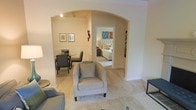This single-floor residence showcases a thoughtfully designed layout that balances comfort and elegance throughout its interconnected living spaces. The living area serves as the heart of the home, furnished with plush seating including a cushioned armchair and coordinating sofa, all set atop soft wall-to-wall carpeting in warm neutral tones.

A defining architectural feature is the graceful arched passageway that elegantly frames the transition between the living area and the adjoining spaces. Through this arch, the dining area is visible to one side, offering an intimate setting with a dark-toned dining table and chairs, complemented by classic wainscoting along the lower walls and framed artwork above. The refined millwork adds a layer of traditional charm to the room.

Beyond the arch, a corridor leads directly to the bedroom, which is appointed with a tufted upholstered headboard and layered bedding in soft, neutral hues. Colorful wall art accents the hallway, adding vibrant personality to the otherwise understated palette.

Throughout the space, the warm beige and cream color scheme creates a cohesive and inviting ambiance. Recessed and track lighting provides focused illumination, enhancing the architectural details and furnishings. The overall design reflects a careful balance of classical detailing and comfortable, livable interiors.