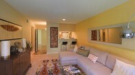 This single-floor residence showcases a thoughtfully designed open-concept layout that seamlessly connects the living area to the kitchen. The living room is anchored by a generously sized sectional sofa and features warm yellow accent walls that contribute to an inviting, cheerful ambiance. A large wall-mounted mirror enhances the sense of space, while recessed lighting and decorative wall art add polished finishing touches throughout.

The kitchen is accessible via a raised breakfast bar with sleek bar stools, creating a natural transition between cooking and living spaces. White appliances and light countertops give the kitchen a clean, bright feel. Carpeted flooring runs throughout the living areas, complemented by a vibrant patterned area rug that adds a pop of color and texture.

A hallway visible from the living room leads to additional areas of the residence, suggesting a practical and well-organized floor plan. The overall aesthetic blends warm, earthy tones with contemporary furnishing choices, creating a comfortable and functional living environment suited for everyday living and casual entertaining.