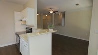 This single-story space showcases an open-concept layout that seamlessly connects the kitchen and living area, creating a bright and cohesive environment. The kitchen is outfitted with white cabinetry and a peninsula topped with a light-toned countertop, offering additional workspace and a natural visual divider between the cooking and living zones. A black electric range is positioned along the kitchen wall, complemented by an overhead ventilation hood. The peninsula provides a convenient casual dining or prep area, with decorative touches such as a small potted plant adding warmth to the space. Transitioning into the living area, rich dark hardwood flooring extends throughout, providing a striking contrast to the neutral, light-toned walls. A ceiling fan with an integrated light fixture is centrally positioned, ensuring air circulation and ambient lighting. Decorative columns frame the transition between the kitchen and living space, adding subtle architectural character. The overall ambiance is clean, functional, and inviting, with a neutral color palette that lends itself well to a variety of interior styles.