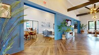 Located at Powell Road in Columbia, SC, the Grand Life Clubhouse is a thoughtfully designed community gathering space that blends modern farmhouse aesthetics with inviting functionality. The single-floor layout encompasses a generously sized open-plan interior highlighted by dramatic vaulted ceilings with exposed dark wood beams, lending the space a warm yet airy atmosphere.

Striking herringbone-patterned hardwood flooring runs throughout the entire clubhouse, providing a cohesive and visually dynamic foundation. Bold blue shiplap accent walls serve as architectural focal points, framing a comfortable lounge area furnished with leather seating, a plush sofa, and a wall-mounted flat-screen TV atop a classic white credenza.

The welcoming reception area features a custom desk branded with "Welcome to the Grand Life," pendant globe chandelier lighting, and natural light filtering through large glass entry doors. Additional seating nooks and a dining area with rattan-style chairs offer flexible spaces for socializing or working.

Lush indoor greenery is thoughtfully placed throughout, enhancing the fresh, vibrant ambiance. Curated abstract wall art, geometric pendant fixtures, and large windows overlooking outdoor greenery complete the polished yet approachable design of this well-appointed clubhouse.