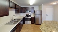 This single-floor residence features a thoughtfully designed layout encompassing one bedroom, one bathroom, and a versatile den. The kitchen serves as a functional centerpiece, outfitted with stainless steel appliances including a refrigerator, range with microwave above, and a double-basin sink. Rich, dark-toned cabinetry provides generous storage, while light granite countertops offer a contrasting visual appeal. Wood-look flooring runs throughout the kitchen and entry areas, adding warmth and durability.

The kitchen opens toward a spacious living area via a granite-topped breakfast bar, creating an open, connected feel. The living area is finished with plush neutral carpeting and features a large window that allows natural light to fill the room. The bedroom mirrors this comfortable aesthetic with carpeted flooring and its own window with blinds.

The bathroom is finished with large-format white ceramic tile flooring, a granite-topped vanity, and clean neutral walls, offering a bright and functional space. The den provides additional flexibility for use as a home office or secondary living area. Throughout the unit, textured ceilings and neutral-toned walls create a cohesive, move-in ready environment.