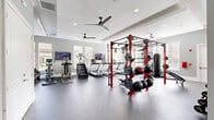 This well-appointed fitness center is designed to support a comprehensive workout experience across multiple training zones. The main exercise area is bright and open, featuring rubber flooring throughout and multiple ceiling fans to maintain air circulation. Large windows with blinds line the walls, allowing generous natural light to fill the space while offering views of the surrounding exterior.

The cardio section is outfitted with a row of treadmills, elliptical trainers, a stair climber, and a rowing machine, each accompanied by wall-mounted flat-screen TVs for entertainment during workouts. A bold red power rack system anchors the strength training zone, complete with barbells, weighted plates, medicine balls, and a heavy punching bag. A rack of dumbbells in graduated weights, kettlebells, and an adjustable weight bench provide versatile free weight options, supported by a cable machine for additional resistance training.

A separate, more intimate training room features a standout warm wood-paneled tray ceiling with a ceiling fan, creating a distinct atmosphere. This space houses additional strength machines and benches, ideal for focused, individual training sessions. A storage unit with cubbies and a wall mirror add practical finishing touches. Throughout the facility, the gray-toned walls and clean architectural details contribute to a polished, professional fitness environment.