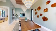 Located at 11590 SW Roma Way in Port Saint Lucie, Florida, this thoughtfully designed single-floor space blends coastal elegance with modern sophistication. The centerpiece of the space is a generously sized dining and meeting room anchored by a long, solid wood farm-style table surrounded by mid-century modern chairs. Overhead, a dramatic tray ceiling clad in rustic wood planks frames a series of cascading tiered chandeliers that serve as a stunning focal point, casting warm, layered light throughout the room. The walls are finished in a calming sage green tone, complemented by curated artwork including large-format photography and a collection of organic wood slice wall sculptures, adding a natural, earthy contrast. A bold herringbone-patterned tile rug in deep blue anchors the dining area, while light-toned floor tiles extend throughout the remainder of the space. A tall arched window with black steel framing invites abundant natural light and frames lush views of swaying palm trees, reinforcing the Florida coastal atmosphere. The adjacent kitchenette is outfitted with warm wood cabinetry, brushed gold hardware, white quartz countertops, a stainless steel refrigerator, and a subway tile backsplash in muted coastal hues, with an additional large window overlooking the tropical surroundings. Together, these spaces create an inviting, resort-inspired environment ideal for gatherings and events.