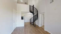 This two-story space makes an immediate impression with its dramatic black spiral staircase, which serves as both a functional element and a bold architectural centerpiece. Soaring double-height ceilings in the main living area amplify the sense of openness and volume, drawing the eye upward and creating an airy, expansive atmosphere throughout the lower level. Warm hardwood-style flooring runs seamlessly across the main floor, lending a cohesive and inviting feel to the space. The open-concept layout connects the living area to a kitchen that features a breakfast bar counter, classic cabinetry, and a chandelier that adds a refined touch overhead. A doorway off the main living area provides access to additional rooms, offering a practical and well-organized floor plan. The upper level, accessible via the spiral staircase, benefits from the height of the vaulted ceiling and overlooks the main living space below. Throughout, the neutral white walls provide a clean, versatile backdrop that complements the contrast of the dark staircase railing and the warm tones of the flooring. This space combines modern design details with functional layout choices, making it both visually distinctive and highly livable.