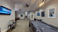 Located at 6800 Gaston Rd in Katy, TX, this single-floor community amenity space is thoughtfully designed to offer residents a versatile and engaging environment for recreation and relaxation. The main game room serves as the centerpiece, featuring a full-size pool table and a shuffleboard table positioned on a stylish patterned carpet that anchors the open layout. Recessed lighting and ceiling fans maintain a comfortable atmosphere throughout, while a wall-mounted flat-screen TV adds an entertainment focal point. Geometric and abstract wall art in navy, gold, and green tones contribute a modern aesthetic to the neutral gray walls. A cozy lounge area offers rich brown leather sofas arranged beneath large windows with white blinds, allowing natural light to filter in and create a warm, inviting ambiance. A dedicated computer workstation alcove provides two desktop stations set against a tiled backsplash wall with pendant lighting overhead, making it a functional space for productivity. The bay window seating area at one end of the game room adds architectural interest and an additional spot for casual gathering. Framed mirrors and curated artwork are distributed throughout, enhancing the polished, community-focused character of this well-rounded amenity space.