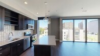 This contemporary urban residence is thoughtfully designed with a clean, modern aesthetic and an open single-floor layout. The expansive living area is anchored by rich dark hardwood flooring and floor-to-ceiling sliding glass doors that flood the space with natural light and frame impressive city skyline views. Globe pendant lights add a stylish focal point to the open-concept kitchen and living zone.

The kitchen is well-appointed with dark espresso cabinetry, light stone countertops, a subway tile backsplash, and a full suite of stainless steel appliances including a refrigerator, electric range, built-in microwave, and dishwasher. A center island provides additional workspace and casual seating.

The bedrooms feature plush carpeting and generous closet space, including a walk-in closet with built-in shelving, hanging rods, and drawers for ample storage. One bedroom benefits from large windows offering additional city views, while the other provides a quieter, more private retreat.

The bathroom is finished with light tile flooring, a dark wood vanity with a single sink and mirrored medicine cabinet, and a glass-enclosed walk-in shower accented with dark tile surround. The overall ambiance of the space is sleek, urban, and move-in ready.