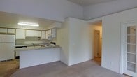 This single-floor residence offers a straightforward and functional layout designed for comfortable everyday living. The open-concept design connects the kitchen and living area seamlessly, creating a cohesive and practical space. The kitchen is equipped with white cabinetry, a built-in sink, and a range, all set beneath under-cabinet fluorescent lighting that ensures a well-lit workspace. A breakfast bar-style counter extends from the kitchen, providing a natural division between the cooking and living zones while maintaining the open feel of the space. The living area features neutral wall-to-wall carpeting that adds warmth and continuity throughout. The ceilings are notably elevated, contributing to a sense of openness and airiness that enhances the overall ambiance. A hallway visible from the main living space leads to additional rooms, suggesting a thoughtful and navigable floor plan. The interior palette is kept neutral throughout, with light-toned walls and finishes that lend the space a clean, bright, and move-in-ready quality. Overall, the layout prioritizes practicality and ease of flow, making it a well-organized and livable single-floor residence.