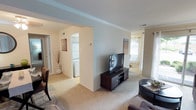 This single-floor residence offers a thoughtfully arranged layout with a warm, neutral aesthetic throughout. The living area serves as the central hub of the home, featuring plush wall-to-wall carpeting, soft light tones on the walls, and elegant crown molding that adds a refined architectural touch to the space. A flat-screen television sits atop a dark wood media console, making this area an inviting spot for relaxation and entertainment. Natural light filters in through a window visible toward the far end of the living space, enhancing the open and airy feel of the room. Decorative column accents contribute subtle architectural character to the space. Adjacent to the living area, a dedicated in-unit laundry space houses a stacked washer and dryer unit, offering added convenience. Passageways connect the various areas of the home, creating a functional and navigable floor plan. Consistent carpeting and a cohesive neutral color palette unify the interior, giving the overall space a clean, move-in-ready appearance. The combination of practical features and comfortable design elements makes this a well-rounded single-level home.