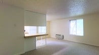 This single-floor space offers a straightforward, functional layout designed to maximize usability. The main living area features wall-to-wall carpeting underfoot and a textured popcorn ceiling overhead, giving the room a classic residential character. Two windows allow natural light to fill the space, creating a bright and open atmosphere throughout the day. A wall-mounted HVAC unit provides convenient climate control directly within the living area.

A defining architectural feature of this space is the pass-through counter that connects the living area to an adjacent room, likely a kitchen or workspace. This built-in counter with a wood-toned surface and metal support legs serves both as a functional workspace and a visual divider, maintaining a sense of openness while subtly delineating separate zones. A roller shade above the pass-through offers privacy and light control between the two areas.

The neutral, cream-toned walls throughout contribute to a clean and airy aesthetic, making the space feel versatile and easy to personalize. The overall design is practical and efficiently organized, suited for everyday living or use as a compact workspace.
