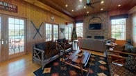 This single-floor lodge-style space exudes a classic cabin ambiance, defined by warm knotty pine wood paneling that lines the walls and ceilings throughout. The open-plan layout connects the living and kitchen areas seamlessly, creating a cohesive and welcoming environment.

The living area is a standout, anchored by a floor-to-ceiling stacked stone fireplace with a wide hearth for storing firewood, and a wall-mounted flat-screen television above. Vaulted wood-planked ceilings with recessed lighting and a ceiling fan add to the airy feel, while large French doors and tall windows flood the room with natural light and offer views of an outdoor deck. Rustic twig-and-branch furniture, Native American-inspired area rugs, and cabin-themed décor — including vintage canoe paddles, snowshoes, and outdoors signage — complete the lodge aesthetic.

The kitchen is well-appointed with warm honey-toned wood cabinetry, dark countertops, and a full suite of stainless steel appliances including a gas range, over-the-range microwave, and a side-by-side refrigerator with ice and water dispenser. A built-in wine rack adds a refined touch. A curved bar peninsula with a wainscoted front and matching dark countertop separates the kitchen from the main living space, accompanied by rustic branch-style bar stools, making it an ideal spot for casual dining or entertaining.