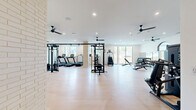 This expansive fitness center offers a generously sized, single-floor layout designed to accommodate a full range of workout activities. The space features light wood-toned flooring throughout, creating a clean and modern aesthetic that complements the crisp white walls and bright, open atmosphere.

Natural light flows in abundantly through large, black-framed windows positioned along multiple walls, providing views of the surrounding outdoors while keeping the interior bright and energizing. Multiple black ceiling fans are distributed evenly across the ceiling, ensuring consistent air circulation throughout the space. Recessed lighting supplements natural light for well-illuminated workouts at any time of day.

The fitness floor is thoughtfully arranged into distinct workout zones. A dedicated cardio area houses multiple treadmills and stationary cycling bikes, along with a rowing machine and elliptical trainer. The strength training section is well-appointed with a multi-station cable machine, a full dumbbell rack with adjustable benches, and a variety of selectorized resistance machines targeting multiple muscle groups. Wall-mounted mirrors are strategically placed to support form checks during exercise.

Architectural details such as an exposed brick column accent and a decorative patterned wall panel add character to the otherwise clean, contemporary design. The overall ambiance is professional, airy, and inviting, making this facility suitable for a comprehensive fitness routine.
