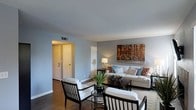 This single-floor space presents a thoughtfully designed living area finished with soft gray walls and rich hardwood flooring that together establish a calm, contemporary ambiance. The living room serves as the central gathering space, furnished with a neutral-toned sofa accented by colorful throw pillows in coral and teal, complemented by matching dark-framed armchairs upholstered in white fabric. A rectangular area rug anchors the seating arrangement around a dark wood coffee table, where a potted tropical plant adds a lively natural element. A console table behind the sofa holds a table lamp that contributes warm ambient lighting. Wall décor includes a large canvas print depicting a vintage bicycle scene, lending the room an artistic focal point. Ceiling-mounted HVAC vents suggest a central climate control system. A hallway visible from the living area leads to additional rooms, with two white paneled doors offering access to adjoining spaces. The overall layout feels open and functional, with a clean, modern aesthetic supported by a consistent neutral color palette and coordinated furnishings throughout.