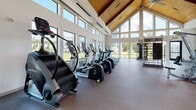 This well-appointed fitness center is designed to inspire an active lifestyle, combining striking architectural details with a full suite of exercise equipment. The space is anchored by a dramatic vaulted ceiling clad in warm wood paneling, which rises to an impressive peak and lends the interior a grand, open atmosphere. Rows of large floor-to-ceiling windows line the walls, flooding the facility with abundant natural light and offering sweeping views of the surrounding outdoor landscape, including palm trees, green lawns, and a resort-style pool area.

The workout floor is covered in durable rubberized flooring and is thoughtfully organized into distinct training zones. A dedicated strength area features a comprehensive rack of dumbbells, weight benches, and multiple cable and selectorized resistance machines. The cardio zone is equipped with a lineup of elliptical trainers, stair climbers, stationary bikes, and rowing machines, all positioned to take full advantage of the scenic window views. Wall-mounted flat-screen televisions are positioned throughout for entertainment during workouts.

Natural light, high ceilings, and open sightlines to the outdoors create an energizing and welcoming ambiance, making this fitness center a standout amenity for health-focused users.