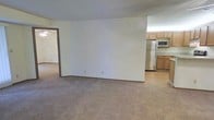 This single-floor residence offers a straightforward, functional layout designed for comfortable everyday living. The main living area is finished with wall-to-wall neutral-toned carpeting, creating a warm and understated atmosphere throughout. Clean white walls keep the space feeling bright and open, providing a versatile backdrop for a variety of interior styles.

A partial wall divides the living area from the kitchen, offering a sense of separation while maintaining an open, connected feel between the two spaces. The kitchen features warm-toned wood cabinetry that extends to the upper walls, providing ample storage. Stainless steel appliances, including a full-size refrigerator and a microwave, lend a modern touch to the kitchen. The kitchen flooring transitions to a smooth hard surface, offering a practical contrast to the carpeted living areas.

A doorway visible from the main living space leads to an additional room, suggesting a multi-room floor plan that accommodates various living needs. The overall ambiance of the space is clean, neutral, and move-in ready, making it well-suited for those looking for a simple and functional home environment. The single-floor layout ensures easy accessibility and seamless flow between living spaces.