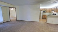 This single-floor residence offers a straightforward, functional layout designed for comfortable everyday living. The main living area is finished with wall-to-wall neutral-toned carpeting, creating a warm and understated atmosphere throughout. Clean white walls keep the space feeling bright and open, providing a versatile backdrop for a variety of interior styles.

A partial wall divides the living area from the kitchen, offering a sense of separation while maintaining an open, connected feel between the two spaces. The kitchen features warm-toned wood cabinetry that extends to the upper walls, providing ample storage. Stainless steel appliances, including a full-size refrigerator and a microwave, lend a modern touch to the kitchen. The kitchen flooring transitions to a smooth hard surface, offering a practical contrast to the carpeted living areas.

A doorway visible from the main living space leads to an additional room, suggesting a multi-room floor plan that accommodates various living needs. The overall ambiance of the space is clean, neutral, and move-in ready, making it well-suited for those looking for a simple and functional home environment. The single-floor layout ensures easy accessibility and seamless flow between living spaces.
