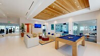 Located at 333 SE Angler Dr in Stuart, Florida, this single-floor amenity center offers a generously sized, thoughtfully designed space that blends social, recreational, and productive environments under one roof. The open-plan layout is anchored by a spacious lounge area featuring neutral sofas and armchairs accented with navy blue pillows, a vibrant tropical-patterned area rug, and warm wood planter boxes filled with ornamental grasses. A wall-mounted flat-screen TV and natural light from large windows and glass doors create an inviting, resort-like atmosphere throughout. A standout wood-slatted ceiling adds warmth and texture, contrasting beautifully with the crisp white walls and large-format tile flooring. The fitness center is enclosed in full-height glass walls, showcasing a well-equipped workout area with a variety of strength and cardio machines, mirrored walls, and direct views to the outdoors. Adjacent to the lounge, a game area features a pool table, while a glass-enclosed co-working suite offers individual workstations with privacy dividers, warm wood flooring, large windows overlooking palm-lined surroundings, and sculptural pendant lighting — ideal for focused work within a stylish setting.