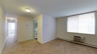 This single-floor residential unit offers a practical and straightforward layout designed for comfortable everyday living. The living area is finished with wall-to-wall carpet in a neutral tone, creating a warm and understated atmosphere. Smooth white walls and a flush ceiling light fixture keep the space feeling clean and bright. A window fitted with horizontal blinds allows natural light to filter into the room, while a wall-mounted air conditioning unit and a baseboard heater provide year-round climate control.

Adjacent to the living area, a separate kitchen is visible through an open doorway. The kitchen is equipped with a white electric range and oven, set against light-colored walls, giving the cooking space a clean and functional character. The transition between the living area and kitchen is defined by a partial wall, offering a degree of separation while maintaining an open connection between the two spaces.

Overall, the unit presents a no-frills, functional living environment with all the essential features in place. The neutral color palette throughout — from the carpeting to the walls and cabinetry — lends the space a cohesive, move-in-ready feel suitable for a wide range of occupants.