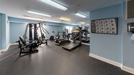 This fitness center is a dedicated, single-floor exercise space designed to support a comprehensive workout routine. The room features soft blue-painted walls that create a calm, focused atmosphere, complemented by durable speckled rubber flooring that provides both safety and comfort underfoot. Overhead fluorescent lighting ensures the space is well-lit throughout.

The facility is outfitted with a varied selection of cardio equipment, including treadmills and an elliptical trainer, positioned along one wall and reflected in large floor-to-ceiling mirrors that visually expand the room and allow users to monitor their form. A multi-station cable and weight machine offers a wide range of strength training options, while additional free weights are available for supplementary exercises.

Natural light filters in through multiple windows fitted with blinds, adding to the open feel of the space. A flat-screen television is mounted on the wall, providing entertainment during workouts. The room also includes a dedicated open-floor area, ideal for stretching, floor exercises, or additional equipment use. A wall-mounted exercise guide chart provides a handy reference for workout routines. The space is accessible via a classic paneled white door and is designed to serve as a fully functional, amenity-style fitness room.