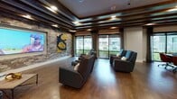 This single-floor amenity space is thoughtfully designed as a multifunctional recreation and social hub, combining entertainment zones with a cohesive industrial-chic aesthetic. Large-format slate-toned tile flooring anchors the billiards area, where a full-size pool table takes center stage beneath warm pendant lighting. A wall-mounted cue rack and high-top bar tables with industrial-style stools create a game-room atmosphere, complemented by bold large-scale portrait artwork and exposed brick accent walls.

Transitioning into the lounge area, rich hardwood flooring and dark exposed beam ceilings lend a warm, intimate character. A wall-mounted flat-screen TV is framed within a stone accent wall, flanked by curated artwork. Comfortable tufted leather sofas and upholstered swivel chairs are arranged for both relaxed viewing and social gathering, with generous windows offering views of the surrounding outdoor greenery and filling the space with natural light.

A dedicated game alcove features a round poker table with leather rolling chairs, alongside a dramatic oversized wall-mounted Scrabble board with an accompanying chalkboard, adding a playful and interactive element. Exposed wooden beam ceilings carry consistently throughout, tying all zones together. The overall ambiance is sophisticated yet inviting, blending upscale finishes with a relaxed, social atmosphere ideal for community engagement.