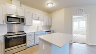 This single-floor unit presents a clean, contemporary design throughout, with a neutral color palette of warm beige tones and crisp white finishes that create a bright and welcoming atmosphere. The kitchen serves as a functional centerpiece, featuring white shaker-style cabinetry, light quartz countertops, a central island with additional prep space, and a full suite of stainless steel appliances including a range, microwave, dishwasher, and refrigerator. Light wood-look flooring runs through the kitchen and main living areas, adding warmth to the space.

The living area is generously proportioned, offering access to a private balcony through sliding glass doors that frame expansive outdoor views and allow abundant natural light to fill the room. The bedrooms feature a mix of light wood flooring and plush carpet, each with windows that provide pleasant views of the surrounding landscape. The bathrooms are finished with white shaker vanities, solid-surface countertops, large mirrors, and brushed nickel hardware. One bathroom includes accessibility grab bars, reflecting a thoughtful and inclusive design approach throughout the unit.